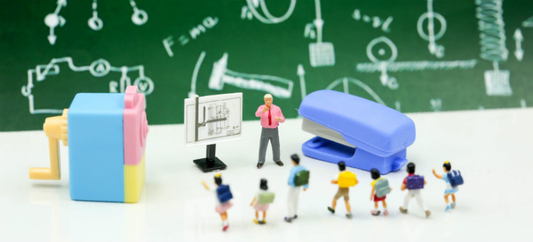 Imagem mostra uma perspectiva de uma mesa com miniaturas de alunos assistindo a um professor, também em miniatura, apresentando dados importantes