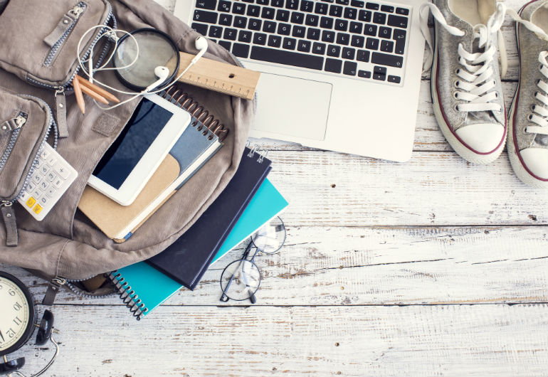 Mochila com tablet, cadernos, lupa sobre uma superfície de madeira. Ao lado, tênis e computador