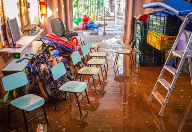 A imagem mostra uma sala inundada com água barrenta. A sala contém cadeiras pequenas, possivelmente de uma escola ou creche, organizadas em fileiras. Brinquedos, bicicletas infantis e equipamentos de playground estão parcialmente submersos na água. Na parede, há uma estante com um monitor de computador e outros objetos. Ao fundo, uma porta aberta revela a área externa. Também há uma escada, algumas caixas de plástico empilhadas e colchões ou almofadas no canto da sala. A retrata danos significativos devido à inundação em Porto Alegre (RS).