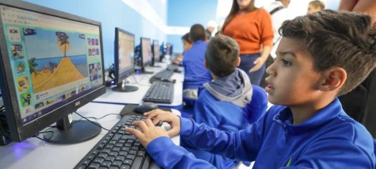 A imagem mostra várias crianças em uma sala de informática usando computadores. Cada uma está concentrada na tela, jogando um jogo educativo colorido. As crianças vestem uniformes azuis. Ao fundo, alguns adultos observam e interagem com elas. A sala é bem iluminada e tem paredes azuis e brancas, dando um ar moderno e acolhedor ao ambiente.