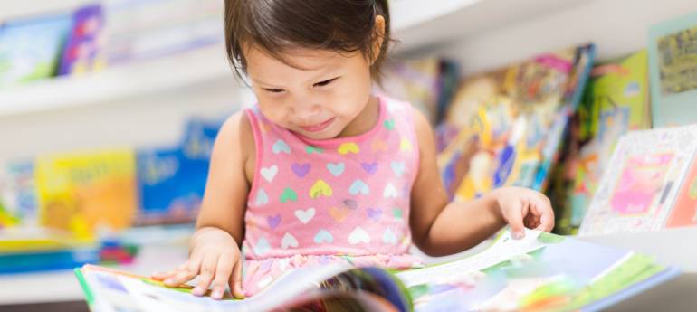A imagem mostra uma criança pequena sorrindo enquanto lê um livro em uma sala de leitura, com estantes de livros ao fundo. A criança está vestida com uma camisa sem mangas rosa, decorada com pequenos corações coloridos. Ela parece estar entretida com o livro que está segurando, enquanto outros livros estão espalhados ao redor.