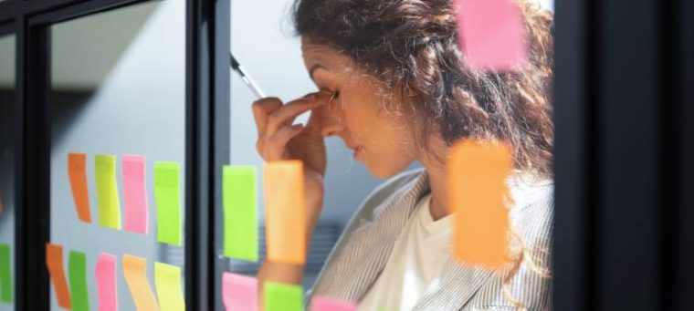 A imagem mostra uma mulher de perfil, com expressão de cansaço, segurando um lápis. Ao redor, há post-its coloridos em um painel de vidro, sugerindo um ambiente de trabalho com muitas tarefas.