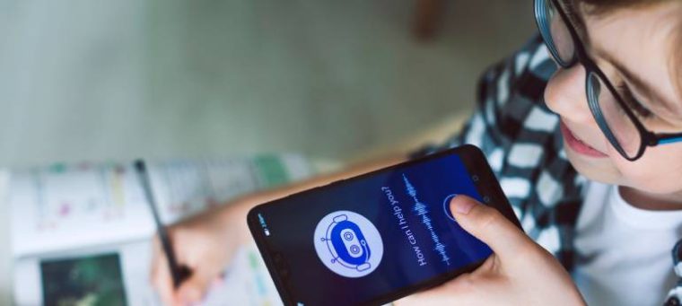 A imagem mostra uma criança com óculos segurando um smartphone com um assistente de voz ativado, exibindo "How can I help you?". Ela escreve em um livro didático enquanto um estojo com lápis de cor está sobre a mesa, sugerindo o uso da tecnologia para auxiliar nos estudos.