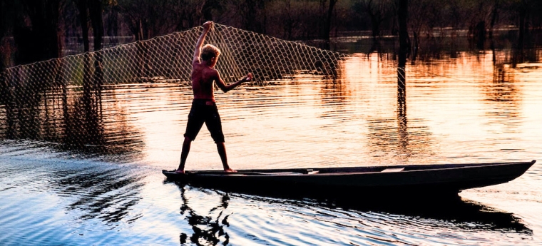 Jovem pescador em uma canoa sobre um rio ao pôr do sol, lançando uma rede de pesca enquanto sua silhueta se reflete na água.