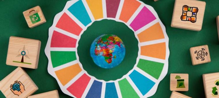Aqui está uma versão reduzida da descrição: A imagem representa os **Objetivos do Desenvolvimento Sustentável (ODS)**, com um círculo colorido ao redor de um pequeno globo terrestre. Blocos de madeira com ícones de sustentabilidade, como reciclagem e energia renovável, cercam a cena. O fundo verde reforça a temática ecológica e a importância dos ODS.