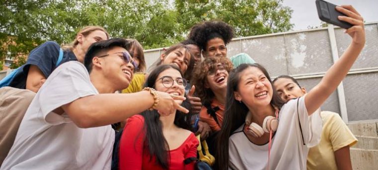 A imagem mostra um grupo de jovens sorrindo enquanto tiram uma selfie ao ar livre, em um ambiente escolar ou público. Eles vestem roupas casuais e coloridas, transmitindo alegria e descontração.