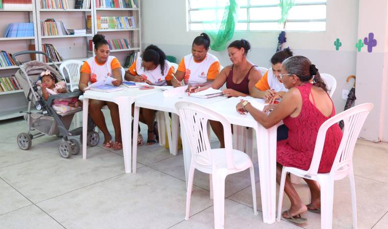 Mulheres adultas estudam em grupo em uma sala de aula, com livros e cadernos. Uma delas cuida de um bebê em carrinho, refletindo os desafios e a força das alunas.