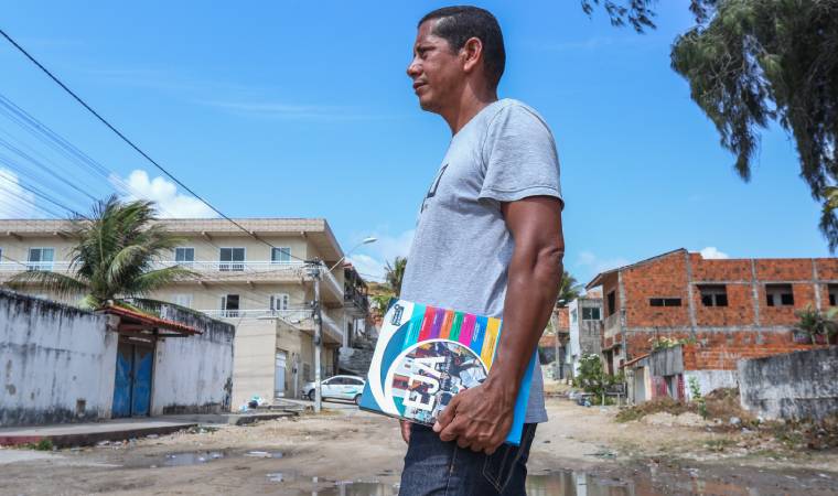 Homem com material da EJA caminha por uma rua simples, mostrando a realidade de quem busca educação em meio a condições adversas