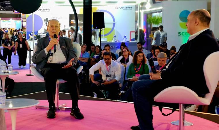 Dois homens em debate em um palco de evento, com plateia e estandes de expositores ao fundo.