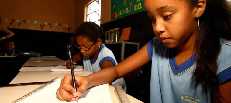 A imagem mostra duas meninas em sala de aula, concentradas em escrever em seus cadernos. Usam uniforme azul. Ao fundo, há cartazes coloridos com temas pedagógicos, indicando um ambiente de aprendizagem acolhedor