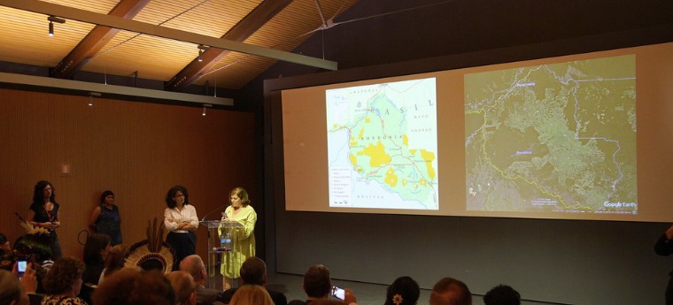 Mulher palestrante fala em púlpito com mapas de Rondônia projetados ao fundo, enquanto público assiste.