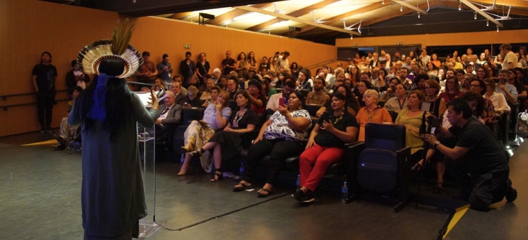 Visão do palco mostra mulher indígena falando para plateia numerosa e atenta em auditório moderno.