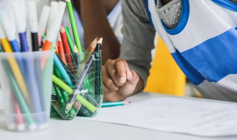 Criança com uniforme escolar desenha ou escreve em uma mesa, ao lado de copos com lápis de cor e canetas.