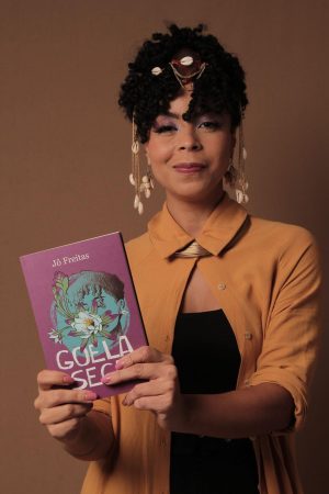 Mulher segurando o livro “Goela Seca”, com acessórios de búzios e roupa mostarda, diante de fundo marrom.