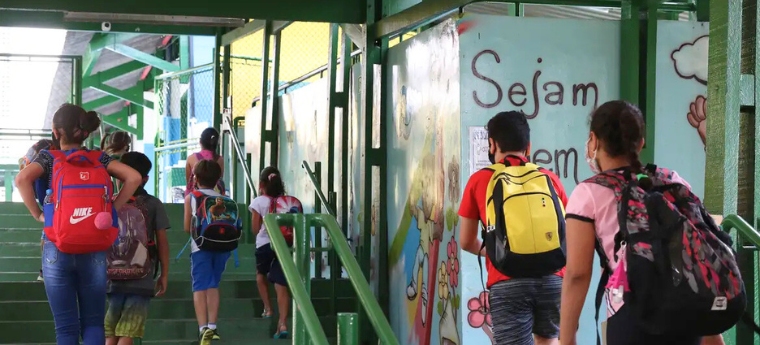 Crianças subindo escada em escola com paredes decoradas e a frase “Sejam bem-vindos”, carregando mochilas.