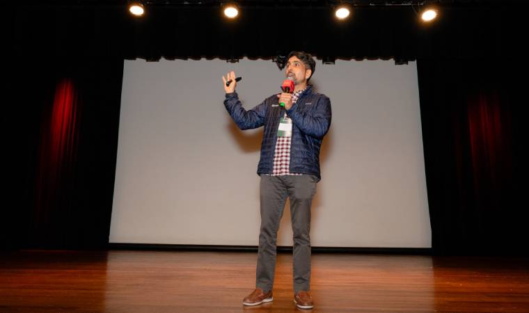 Sal Khan fazendo apresentação em um palco com microfone e controle, em frente a uma tela vazia