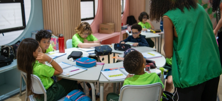 Sala de aula com crianças fazendo atividades e professora acompanhando.