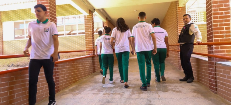 Estudantes caminham em um corredor escolar, enquanto um segurança observa encostado na lateral.