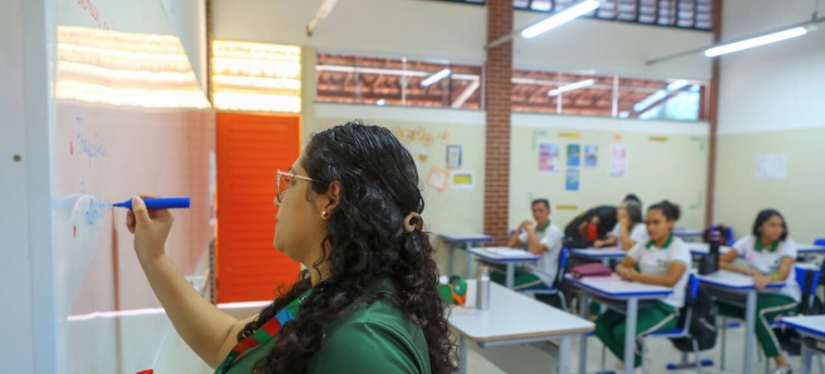 Professora escreve no quadro enquanto alunos, sentados nas carteiras, acompanham a aula.