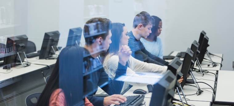 Grupo de cinco pessoas em uma sala de informática, sentadas diante de computadores e digitando. A imagem é refletida através de uma janela transparente, criando um efeito de sobreposição.
