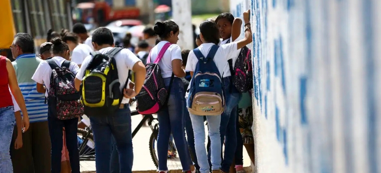 Grupo de estudantes com mochilas reunidos na calçada em frente a um muro, próximo à entrada de uma escola.