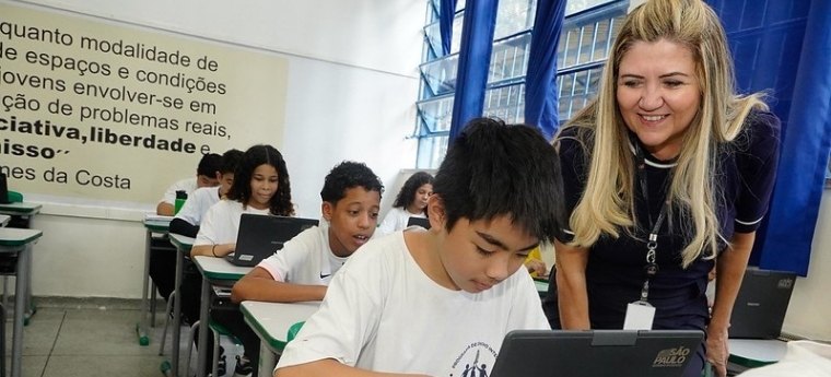 Alunos usando notebooks em sala de aula enquanto professora observa com sorriso.