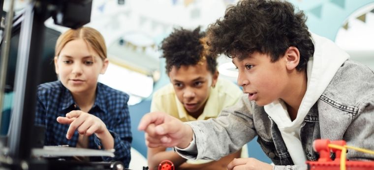 Três adolescentes interagem com impressora 3D em atividade prática de tecnologia.