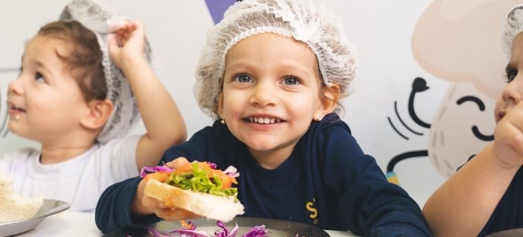 Criança segura um sanduíche com vegetais durante atividade de alimentação saudável em grupo.