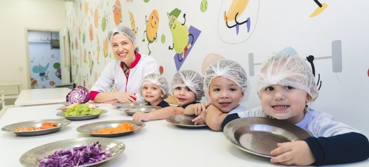 Crianças e educadora sentadas à mesa com alimentos coloridos, em atividade sobre alimentação saudável.
