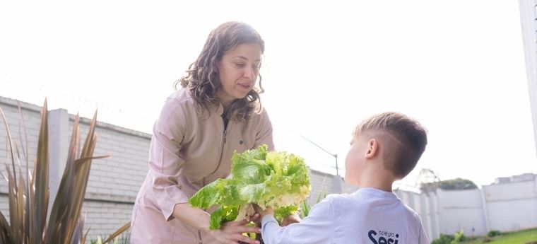 Educadora entrega pé de alface para criança em atividade na horta escolar.
