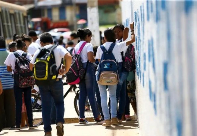 Grupo de estudantes com mochilas conversando em frente a um muro pintado, próximo a um ônibus escolar.