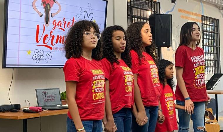 Grupo de meninas em apresentação sobre autoestima e educação corporal, diante de tela com a frase 'Garotas Vermelhas'.