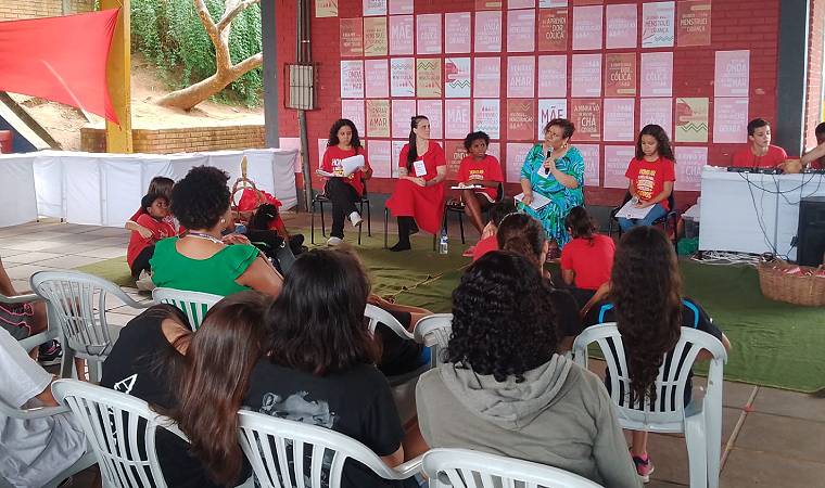 Debate com mulheres e jovens sobre temas sociais e educação corporal, com público atento e cartazes ao fundo.