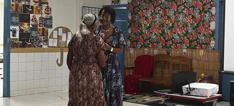 Duas mulheres interagindo em uma sala com decoração floral. Uma mulher está de costas, com cabelo grisalho e vestido marrom. A outra mulher, com cabelo curto e vestido azul floral, está conversando com ela.