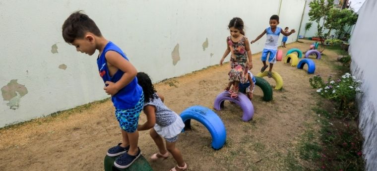 Crianças brincam em uma trilha de pneus coloridos dispostos no chão entre dois muros, em um pátio escolar.