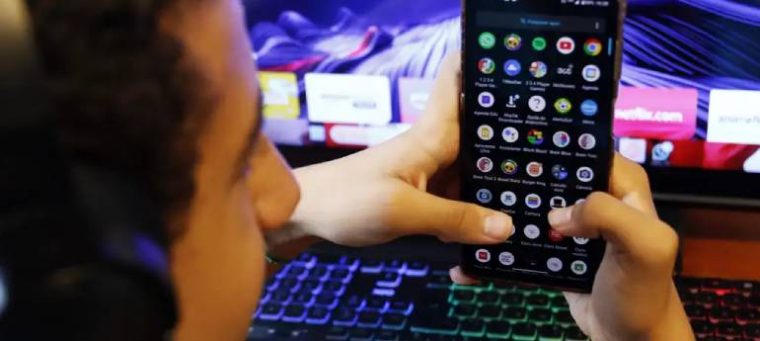 A imagem mostra um jovem segurando um smartphone, com a tela exibindo aplicativos, enquanto está sentado diante de um computador com um teclado iluminado. Ele usa fones de ouvido, sugerindo estar envolvido em uma atividade digital.