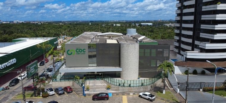 Fachada do prédio da escola COC São Luís, com estrutura moderna, torre cilíndrica central e logomarca visível. Ao lado, um supermercado e um prédio residencial.