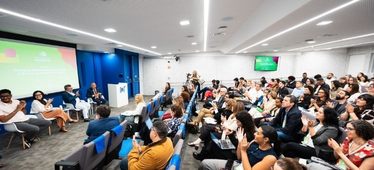 Público aplaude painel em auditório moderno com cinco participantes.