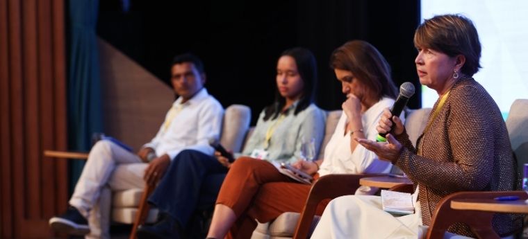 Quatro pessoas em um painel de discussão; uma mulher fala ao microfone enquanto os demais escutam.