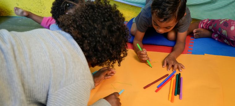 Crianças desenham com canetinhas sobre papel em espaço infantil acolchoado.