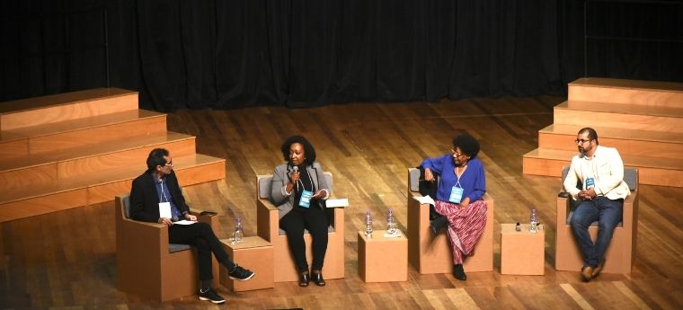 Painel com quatro pessoas sentadas em palco de madeira durante uma conferência, participando de um debate.
