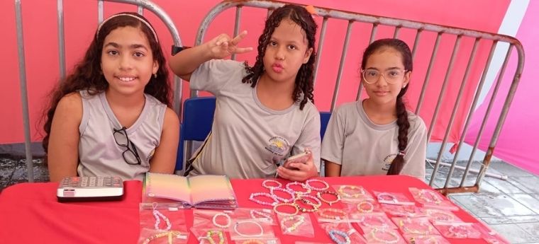 Três meninas expõem pulseiras artesanais em banca da feira escolar de jovens empreendedores.