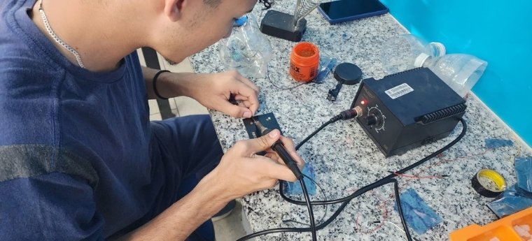 Jovem usando ferro de solda em bancada com equipamentos eletrônicos.