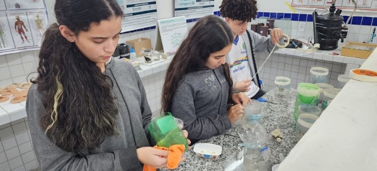 Estudantes manipulando garrafas plásticas e materiais recicláveis em bancada de laboratório escolar.