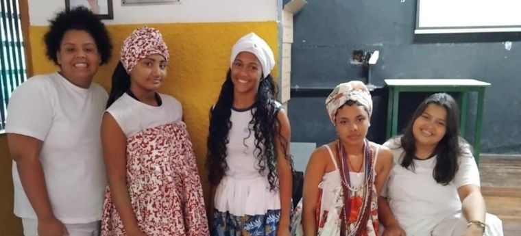 Cinco meninas em ambiente escolar; algumas usam trajes típicos de religiões afro-brasileiras com turbantes e colares, outras vestem branco simples.