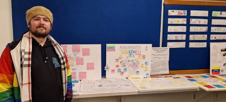 Homem com barba e gorro bege, usando casaco colorido com as cores do arco-íris, posando ao lado de uma mesa com cartazes e maquetes sobre a Venezuela. Ao fundo, painel azul com informações e desenhos sobre a Venezuela e seus países vizinhos.