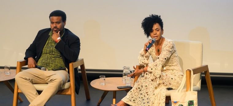 Homem e mulher sentados em palco; ela fala ao microfone enquanto ele observa pensativo.