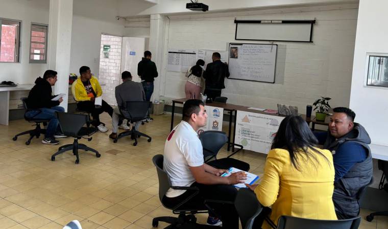 Sala com adultos em atendimento educacional, sentados em duplas e interagindo com materiais informativos nas paredes.