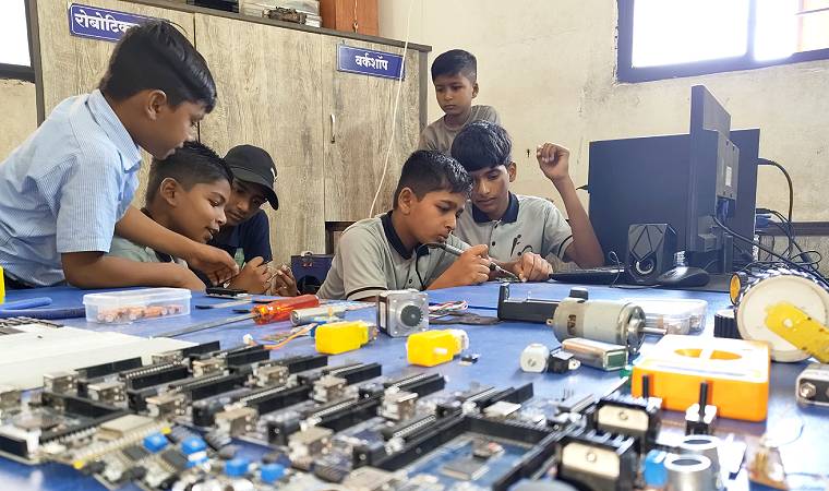 Alunos uniformizados manuseiam peças de robótica em mesa de aula, com foco em atividade prática de eletrônica.