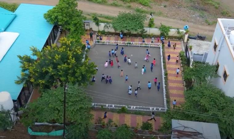 Foto aérea de pátio escolar com crianças brincando em grupos, cercado por árvores e edifícios.